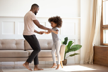 Happy African family father and cute little kid daughter jump having fun in living room in the...