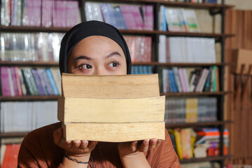 Portrait of Asian hijab woman covering her head with a pile of books in front of library bookshelf....