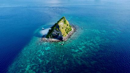 Aerial view of a small rocky uninhabited island in the center of the ocean. Top view of an underwater coral reef around a small tropical island in the open sea, a small sandy beach and a motor boat.