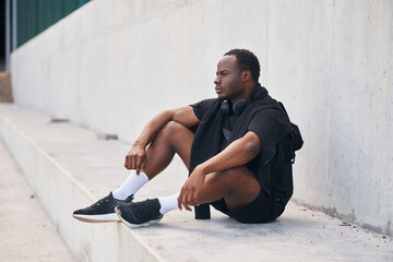 Sitting by the white wall. Handsome black man is posing outdoors at daytime