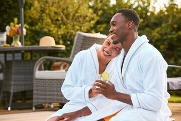 Loving Couple Wearing Robes Outdoors Sitting With Drinks By Swimming Pool On Spa Day