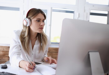 Beautiful woman wearing headphones making notes with silver pen while working on computer pc...