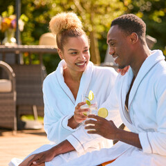 Couple Wearing Robes Outdoors Sitting Doing Cheers With Drinks By Swimming Pool On Spa Day