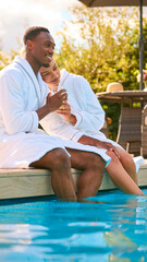 Fototapeta na wymiar Couple Wearing Robes Outdoors Sitting With Drinks With Feet In Swimming Pool On Spa Day