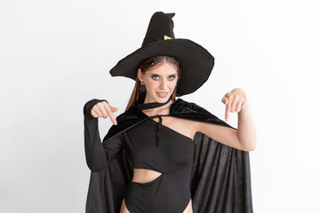 Attractive young woman in black clothes and black hat points her fingers down. White background. Halloween party.