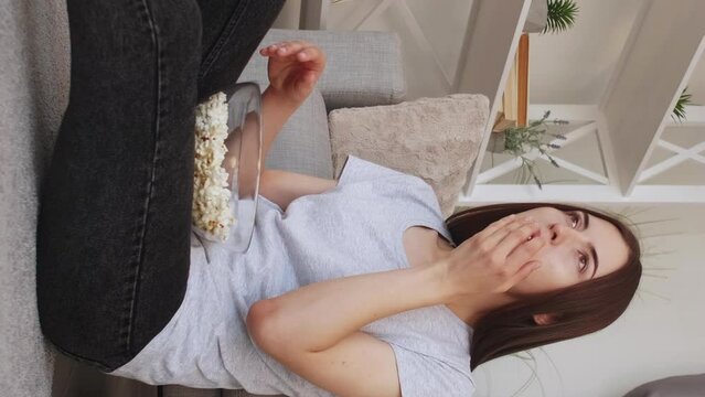 Vertical video. TV cheering. Home leisure. Excited woman. Happy joyful girl eating pop-corn expressing win clenched fists applauding sitting on floor in light room interior.
