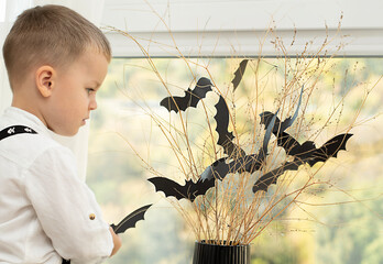 Halloween concept. Autumn holiday. The boy stands with his back near the window against the backdrop of a black vase with batting mice cut out of paper on dry branches. Close-up.