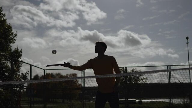 Silhouette of man on fenced playground playing pickleball and hitting ball with racket. Warming up for coordination on outdoor court before pickleball match against cloudy sky.