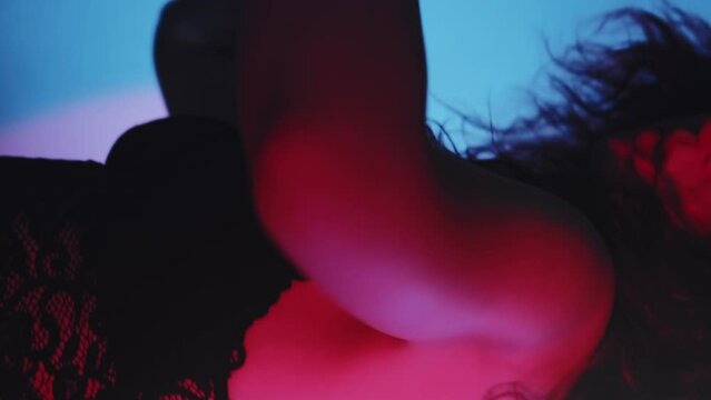 Vertical video. Lingerie dance. Femininity energy. Graceful attractive woman in black lace bralette moving on blur haze neon light background.