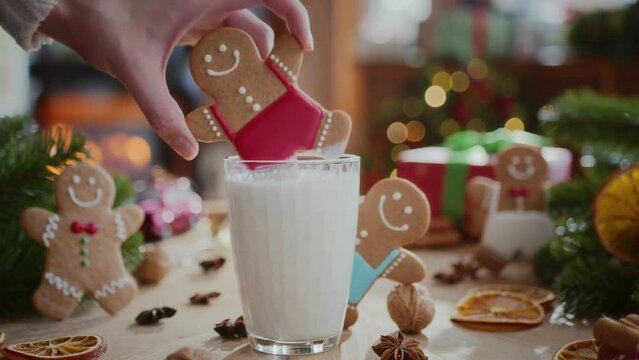 cozy composition of christmas decoration in daylight. woman hand dips gingerbread man cookie in glass of warm milk on wooden table with presents, flashing lights and burning fireplace on background