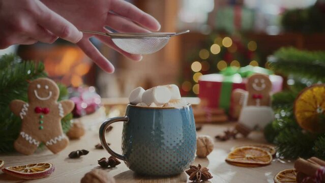 cozy composition of christmas. sprinkling cinnamon spices on marshmallows in blue cup with cocoa on table with fir tree branches, presents and new year decorations, flame in fireplace