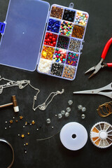 Box with colorful beads, string, wire, chain, scissors, pliers and hammer on dark background. Various jewelry making supplies. Top view.