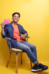 Full length of excited young man sitting on armchair and showing comment sign with happy expression