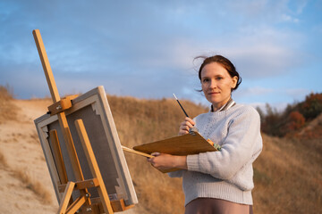 A young woman stands on a beach at sunset and paints an oil painting on an easel. The artist paints a picture of the river bank.