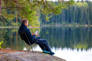 A man reads an e-book on the shore of a lake.