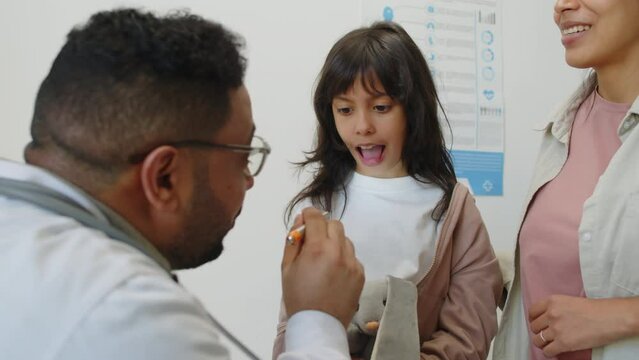 Middle Eastern male child otolaryngologist examining throat of Hispanic little girl with flashlight and giving recommendations to girls mom