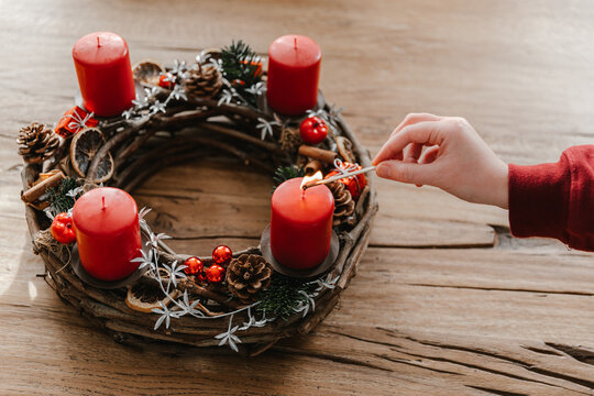 First candle on advent wreath made with branches. Human hand holding a matchstick.