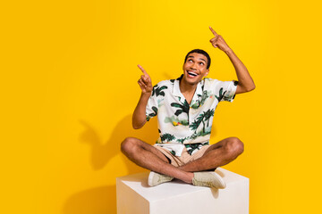 Full body photo of attractive young man sit cube point look empty space wear trendy palms print clothes isolated on yellow color background