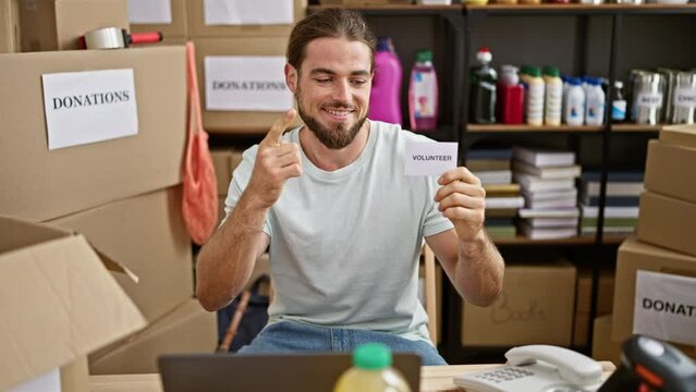 Young hispanic man holding volunteer paper pointing to camera at charity center