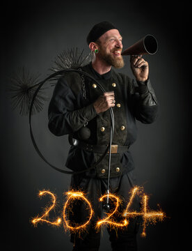 Schornsteinfeger in Arbeitskleidung mit Flüstertüte in der Hand, Jahreszahlen 2024 mit Wunderkerzen geschrieben, Symbolbild für Jahreswechsel, Studioaufnahme.