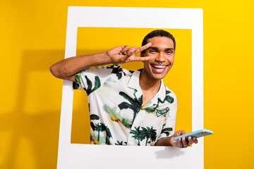 Photo of good mood man with nose piercing dressed shirt hold smartphone show v-sign posing in frame isolated on yellow color background