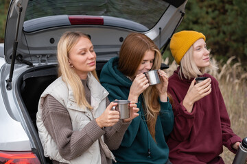 Happy girls sit in the trunk of the car and drink tea from a thermos. Friends traveling by car in...