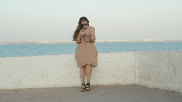 A Young Woman on a Seaside Pier retrieves her Phone from her Skirt Pocket and looks at it while adjusting her Hair, in Slow Motion.