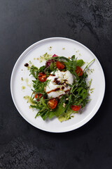 Salad with arugula, tomato and burrata cheese on a dark background.