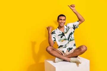 Full size photo of handsome young guy sit white podium point empty space wear trendy palms print outfit isolated on yellow color background