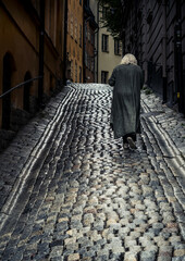 Eine ältere Frau mit weißen längeren Haaren geht gebückt eine regennasse Straße nach oben, das...