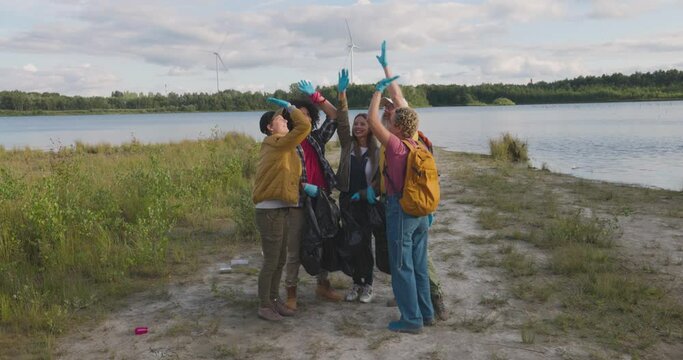 Handsome hardworking multiracial men and women millennials helping to collect rubbish into plastic bags with their friends, nature activists and giving high five each other ,society against pollution