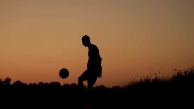 Full length video capturing a silhouette of a teenager, young man playing with a ball on a field at sunset. He is kicking the ball in the air.
