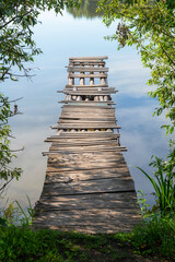 A wooden bridge made of logs and partially rotten boards. Being careful is dangerous. Water, lake, bush. Shore.