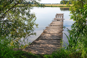 A wooden bridge made of logs and partially rotten boards. Being careful is dangerous. Water, lake, bush. Shore.