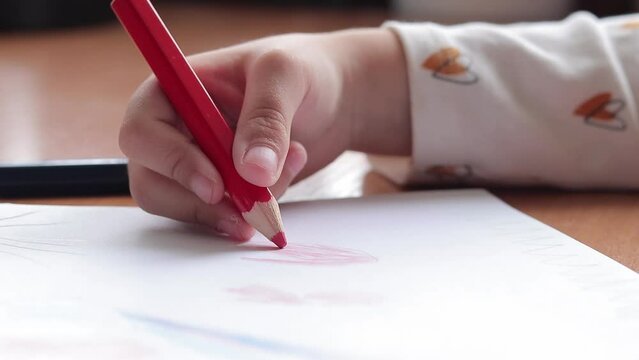 A preschool child draws with colored pencils in an album. Child's hand with a pencil close-up, selective focus. The girl draws hearts in red
