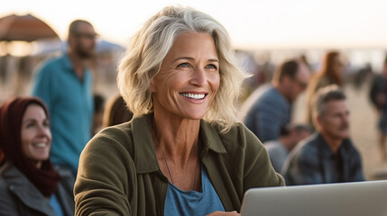 An older white woman smiles while using a laptop on a beach bench, embracing outdoor work or leisure.