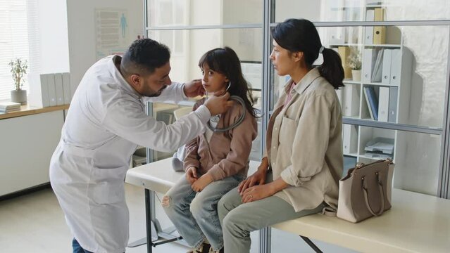 Medium shot of Middle Eastern male pediatrician giving stethoscope to Hispanic little girl so she could check her heartbeat during checkup appointment in modern doctor office