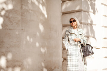 Obraz premium A beauttiful woman with grey hair is walking arounв the old town. A stylidh woman is wearing a gray coat, a skirt and a bag and sunglasses