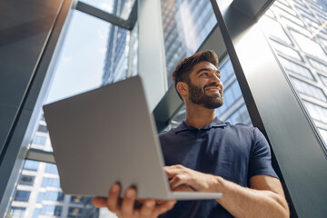 Handsome freelancer with laptop standing in coworking near window and looks away