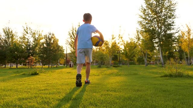 Silhouette of a pine child go on green grass in a park. A boy walking along a meadow holding a ball in his hand. The kid is a volleyball player. dreaming of becoming professional player