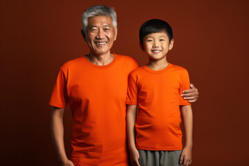 Fototapeta premium Generations United: Smiling Asian Grandpa and Grandson, Both in Orange Shirts, Stand Side by Side