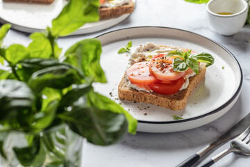 Sandwich or toast with tomatoes, cream cheese, olive oil and basil on a plate on white marble background. Traditional italian mediterranean food