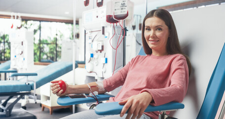 Caucasian Woman Donating Blood For People In Need In Hospital. Female Donor Squeezing Heart Shaped Ball To Pump Blood. Looking Into Camera And Smiling. Donation Center For Victims Of Accidents. - obrazy, fototapety, plakaty