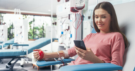 Caucasian Woman Donating Blood For People In Need. Female Donor Squeezing Heart-Shaped Red Ball To Pump Blood And Chatting Online Using Smartphone. Donation For Women With Pregnancy Complications. - obrazy, fototapety, plakaty