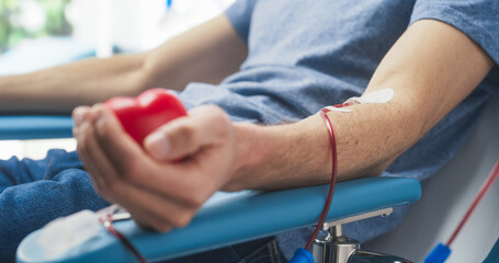 Close Up Shot Of Hand Of Male Blood Donor With an Attached Catheter. Caucasian Man Squeezing Heart-Shaped Red Ball To Pump Blood Through The Tubing Into Bag. Donation For Victims Of Accidents. - obrazy, fototapety, plakaty