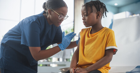 Young African American Boy Sitting In The Chair In Bright Hospital And Getting His Polio Vaccine. Female Black Nurse Is Performing Injection. Professional Woman Talks To Worried Kid And Calms Him Down - obrazy, fototapety, plakaty