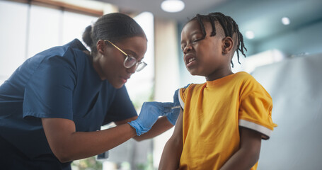 Young African American Boy Sitting In The Chair In Bright Hospital And Getting His Flu Vaccine. Female Black Nurse Is Performing Injection. Professional Woman Being Careful and Gentle To Ease The Pain - obrazy, fototapety, plakaty