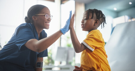 Young African American Boy Sitting In The Chair In Bright Hospital And Getting His Flu Vaccine. Female Black Nurse Is Finished Performing Injection. Professional Woman High-Fives A Kid For Being Brave - obrazy, fototapety, plakaty