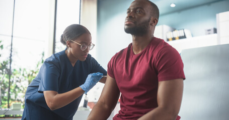Happy African Man Sitting In The Chair In Bright Hospital And Getting His Hepatitis B Vaccine. Professional Black Female Nurse Is Performing Injection And Putting A Patch On. Public Healthcare Concept - obrazy, fototapety, plakaty