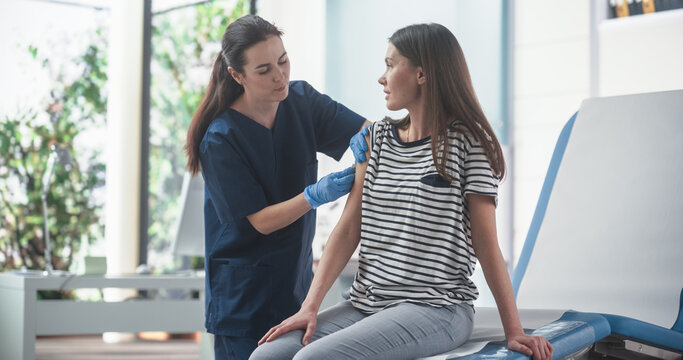 Happy Caucasian Woman Sitting In The Chair In Bright Hospital And Getting Her MMR Vaccine. Professional Female Nurse Is Performing Injection And Putting A Patch On. Public Healthcare Concept.